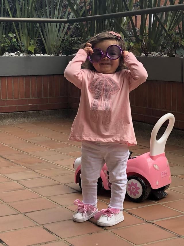 A young child wearing oversized purple sunglasses, smiling beside her pink ride-on car